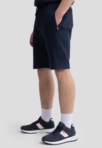 Shorts bleu marine en tissu doux, dotés d'une poche zippée et d'une taille à cordon. Associés à des chaussures de sport bleu marine avec des accents rouges.