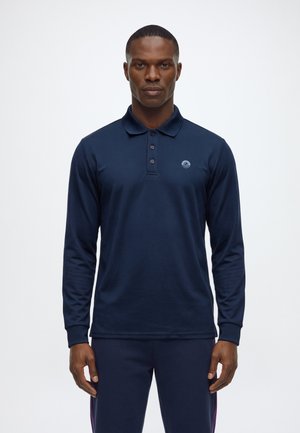 Homme portant un polo bleu marine à manches longues avec des boutons et un logo discret sur la poitrine gauche, debout devant un fond blanc uni.