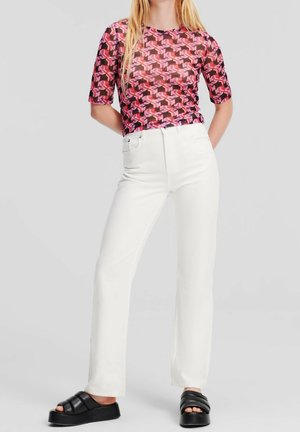 Vrouw die een roze patroon top draagt, nauwsluitend, hoge taille witte rechte jeans en zwarte platformsandalen, staand met handen achter de rug.