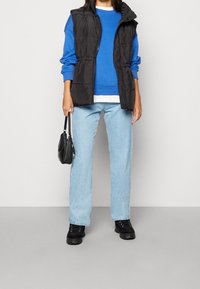 Czarna kamizelka puchowa na niebieskiej bluzie z białymi detalami. Jasnoniebieskie jeansy typu straight-leg i czarne, masywne trampki. Mała czarna torebka.