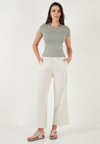 Vrouw staat met haar handen in de zakken, gekleed in een aansluitend grijs T-shirt met korte mouwen, losse witte broek en witte sandalen met decoratieve stenen.