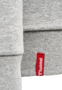 Szary materiał bluzy z gładką teksturą. Posiada czerwoną metkę z białym napisem "hummel" na dolnej części. Prosty, praktyczny design.
