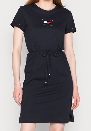 Kobieta w czarnej sukience z krótkim rękawem, ze sznurkiem w talii i logo Tommy Hilfiger na piersi.