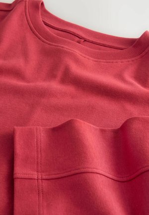 Röd sweatshirt med rundad halsringning, som har ett texturerat tyg, synlig sömdetalj och en något förhöjd ficksektion.