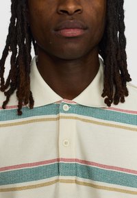 Polo-shirt in crème met een textuurafwerking, met horizontale strepen in groen, rood en geel. Drieknoopssluiting bij de hals.