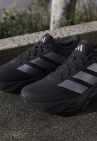 Ζευγάρι μαύρα αθλητικά παπούτσια Adidas με γκρι ρίγες που ακουμπούν σε πλακόστρωτο κοντά σε πεζούλι από πέτρα και φυτά στο παρασκήνιο.