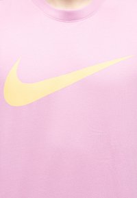 Κοντινό πλάνο του κίτρινου λογότυπου Nike Swoosh τυπωμένου σε ανοιχτό ροζ μπλουζάκι με στρογγυλή λαιμόκοψη.