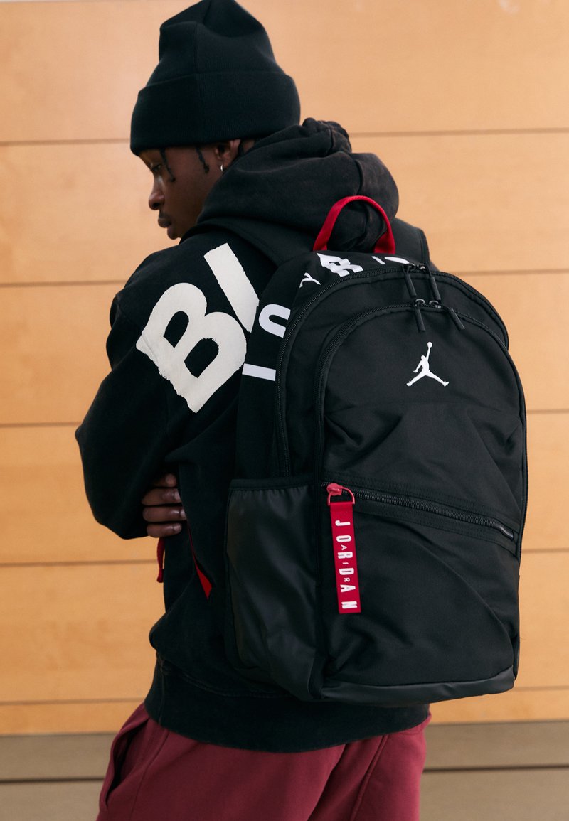 Fekete hátizsák piros díszítéssel, kiemelkedő logóval, cipzáras zsebekkel és texturált szövettel. Egy kapucnis pulóvert viselő személy hátán viselve.