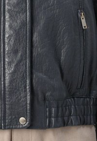 Giacca in pelle nera con una finitura testurizzata, caratterizzata da una cerniera argentata e dettagli con bottoni a scatto. La vita a coste aggiunge un design sottile.