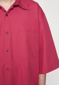 Korte mouwen shirt in een dieprode stof met een puntige kraag, voorzak en knoopsluiting. Gladde textuur, minimalistisch ontwerp.