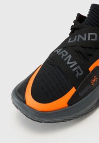 Pantof de sport negru cu un accent portocaliu, un superior texturat, benzi elastice cu branding și o talpă din cauciuc gri.