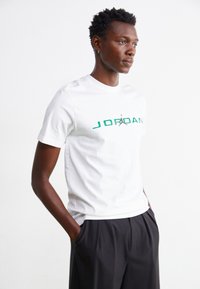 Hvit T-skjorte med korte ermer som har grønn "JORDAN" tekst og logo i sentrum. Stoffet er glat og sitter komfortabelt rundt torsoen.