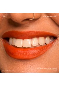 Close-up of a smiling mouth with bright orange matte lipstick and a silver nose ring on warm brown skin.