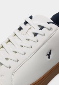 Λευκό αθλητικό παπούτσι με λευκούς κορδόνια, εσωτερική επένδυση σε ναυτικό μπλε, λογότυπο πουλιού σε ναυτικό μπλε στην πλευρά και καφέ ελαστική σόλα, εμφανιζόμενο σε κοντινή λήψη.