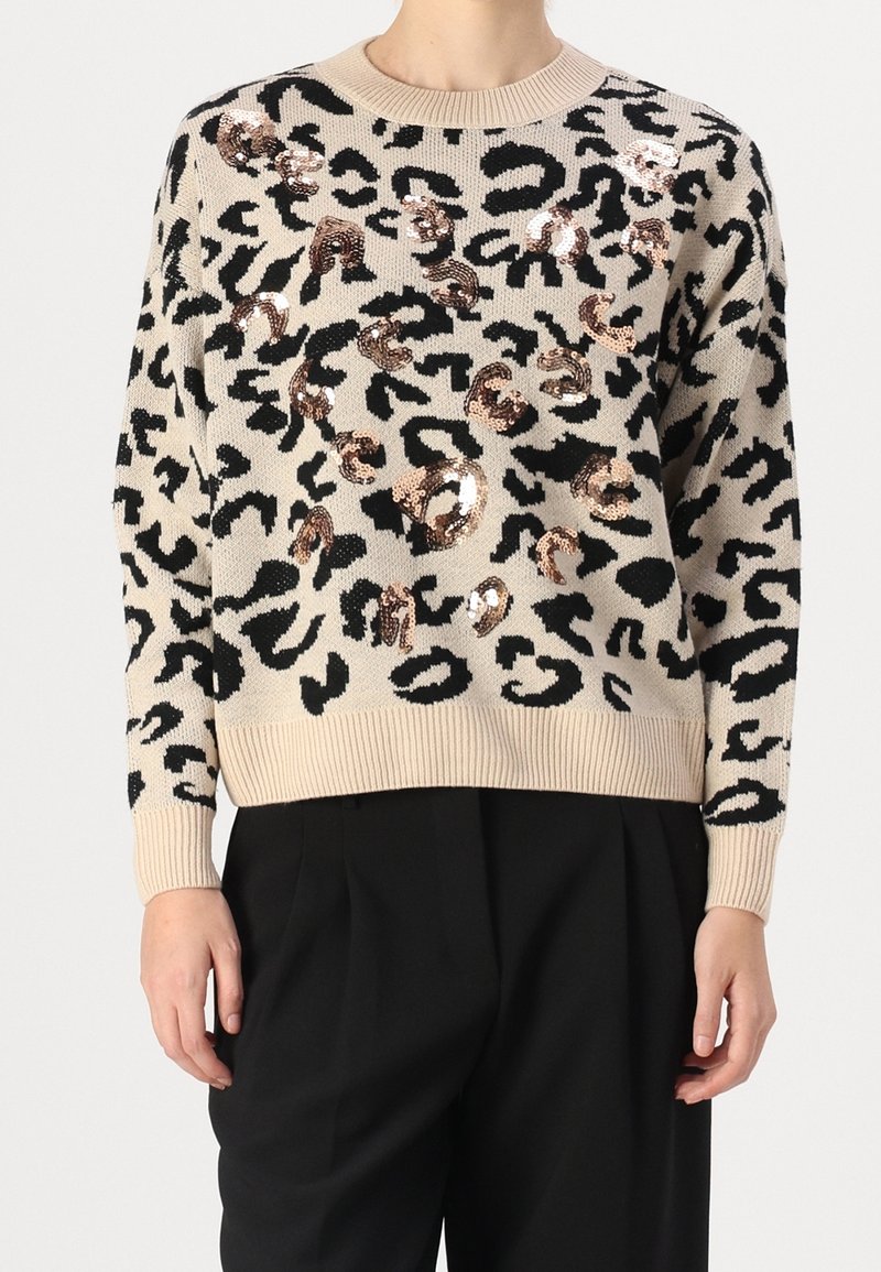 Pull beige avec imprimé léopard noir et accents de sequins bronze brillants, porté avec un pantalon noir.