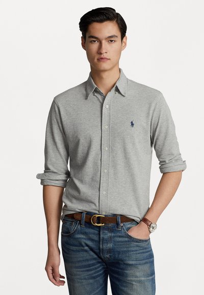 Helles graues Langarmhemd mit Button-Down-Kragen und hochgekrempelten Ärmeln, versehen mit einem kleinen navyblauen Logo, kombiniert mit dunkelblauen Jeans und einem braunen Gürtel.