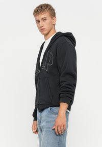 Ung man med kort blont hår som bär en svart zip-up hoodie och ljusblå jeans, stående mot en vit bakgrund.