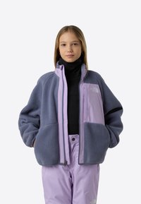 Jopica iz flisa v temno sivi in sivolila barvi, z zadrgo, visokim ovratnikom in logotipom. Oblečena čez črni puli in sivolila hlače.