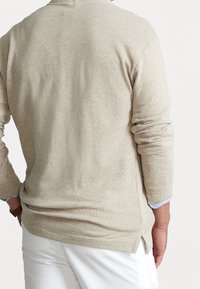 Pull en tricot beige avec des manches longues, fentes sur les côtés à l'ourlet et finition texturée. Porté sur un pantalon de couleur claire.