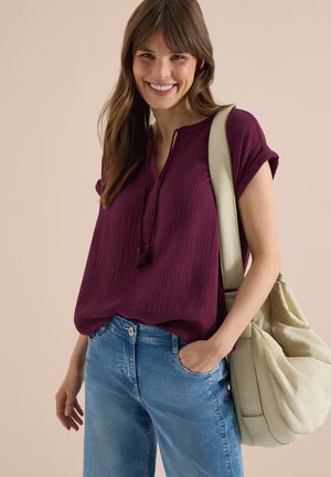 Femme souriante portant un haut violet à manches courtes et un jean bleu, portant un grand sac à épaule beige, main dans la poche.