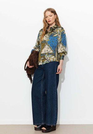 Femme portant un chemisier à motifs bleus et verts, jean large foncé, sandales compensées, tenant un sac marron à franges sur un fond blanc.