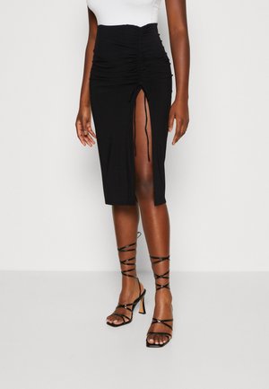 Frau trägt einen schwarzen, gerafften Midirock mit vorderem Schlitz und schwarze, mit Riemchen versehene High-Heel-Sandalen vor weißem Hintergrund.