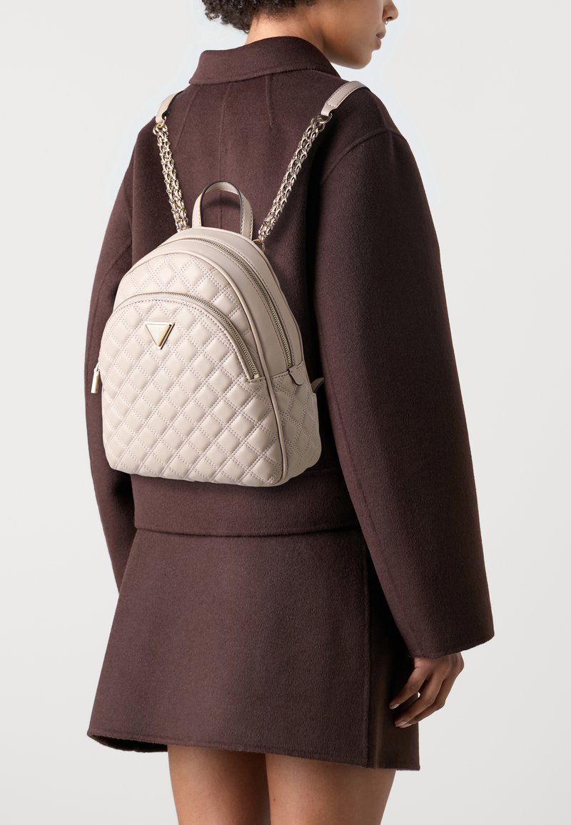 Hellegrauer, gesteppten Rucksack mit doppeltem Reißverschluss, ketteverzierten Trägern und einem dreieckigen Logo. Getragen über einem dunkelbraunen Mantel.