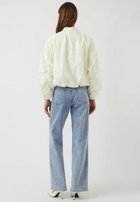 Witte, gestructureerde bomberjack met bloemmotief en volumineuze mouwen, gecombineerd met lichtblauwe, rechte jeans en witte puntige hakken.
