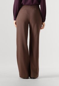 Bruine, wijduitlopende broek met een gladde textuur, voorzien van twee achterzakken en een getailleerde tailleband. Gecombineerd met een paars shirt met lange mouwen.