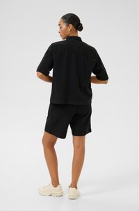 Black linen short-sleeve shirt paired with black shorts. The outfit features a relaxed fit and soft texture. Light-colored sneakers complete the look.