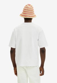 Biała bawełniana koszulka z krótkim rękawem, okrągłym dekoltem i luźnym krojem. W połączeniu z wielokolorową tkanymi kapeluszem typu bucket.