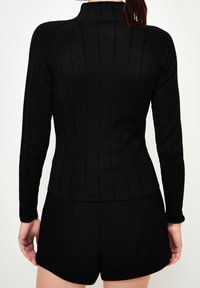 Woman facing away wearing a fitted black long-sleeve ribbed top and black cuffed shorts against a plain light background.