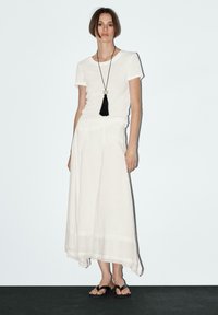 T-shirt bianco, gonna bianca fluida con orlo testurizzato, collana con frange nera e sandali neri, il tutto su uno sfondo chiaro.