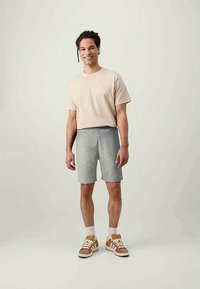 Béžové bavlnené tričko a šedé úpletové šortky, kombinované s bielymi ponožkami a hnedými teniskami. Model stojí proti neutrálnemu pozadiu.