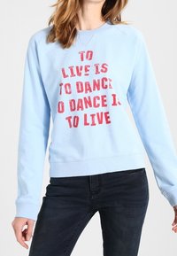 Osoba v světlé modré mikině s červeným nápisem "ŽÍT JE TANČIT, TANČIT JE ŽÍT" a tmavých džínách, stojící na bílém pozadí.