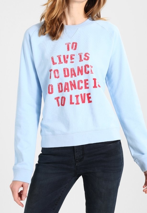 Osoba v světlé modré mikině s červeným nápisem "ŽÍT JE TANČIT, TANČIT JE ŽÍT" a tmavých džínách, stojící na bílém pozadí.