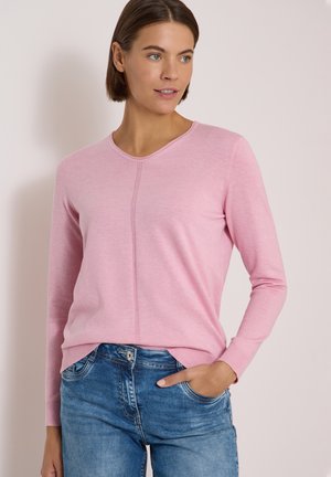 Femme aux cheveux courts bruns portant un haut à manches longues rose clair et un jean bleu, debout avec une main dans la poche devant un fond uni.
