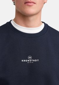 Mørkeblå genser med rund hals laget av mykt stoff, med en hvit logo og teksten "KRONSTADT" og "CASUAL SINCE 1999" på brystet.