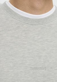 Sweatshirt gris clair avec une finition texturée, présentant un col rond blanc et un logo de la marque "Selected" subtilement embossé sur la poitrine.