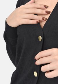 Cardigan nero con tessuto strutturato e scollo a V, chiuso con un bottone rotondo dorato. Le mani sono appoggiate sul capo, mostrando l'arte delle unghie.