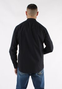 Maglietta nera a maniche lunghe con taglio dritto, polsini con bottoni e colletto alto; indossata con jeans in denim blu con tasche posteriori decorative.
