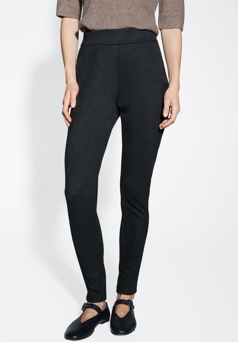 Leggings neri elasticizzati con design a vita alta. Texture liscia senza chiusure visibili. Abbinati a scarpe nere basse e un maglione marrone.