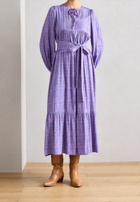 Lilla blomsterkjole med lange ærmer, rynket halsudskæring, taljebånd og udvidet nederdel. Kombineret med beige cowboystøvler.