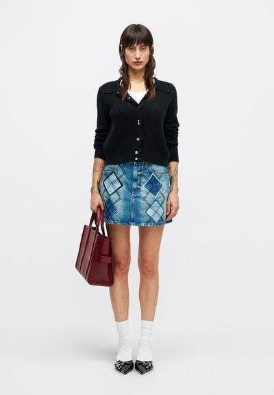 Ženska stoji ob beli steni, oblečena v črn kardigan, modro krilo iz kvadratnih kosov denima, bele nogavice, črne špičaste čevlje in drži bordo torbico.