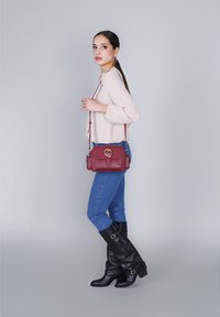 Borsa a tracolla bordeaux in materiale strutturato con chiusura ad anello dorato, tracolla rimovibile e pattina anteriore, abbinata a jeans blu e stivali neri.