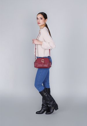 Borsa a tracolla bordeaux in materiale strutturato con chiusura ad anello dorato, tracolla rimovibile e pattina anteriore, abbinata a jeans blu e stivali neri.