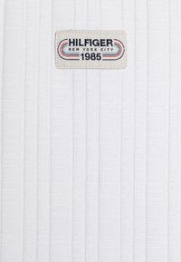 Biała prążkowana tkanina z na przemian pojawiającymi się teksturowanymi wzorami. Tkana metka przedstawia napis "HILFIGER NEW YORK CITY 1985" w niebieskich i czerwonych literach.