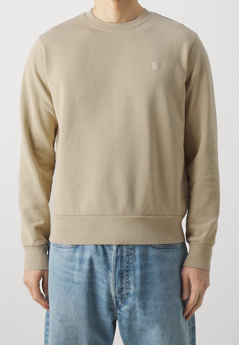 Homme portant un sweat-shirt beige à col rond avec un petit logo brodé sur la poitrine et un jean en denim bleu clair, debout devant un fond uni.