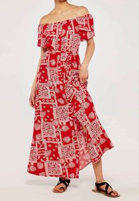 Robe longue rouge avec manches épaules dénudées, motifs floraux et géométriques blancs, détail noué à la taille et fente latérale, portée avec des sandales plates.