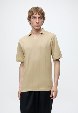 Homme blond portant une chemise beige à manches courtes avec de fines rayures verticales et un pantalon noir, debout sur fond blanc.